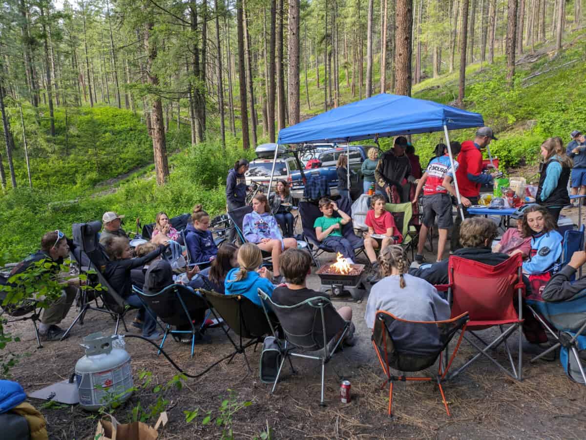 Team members and families eating dinner at campsite and sitting around fire.